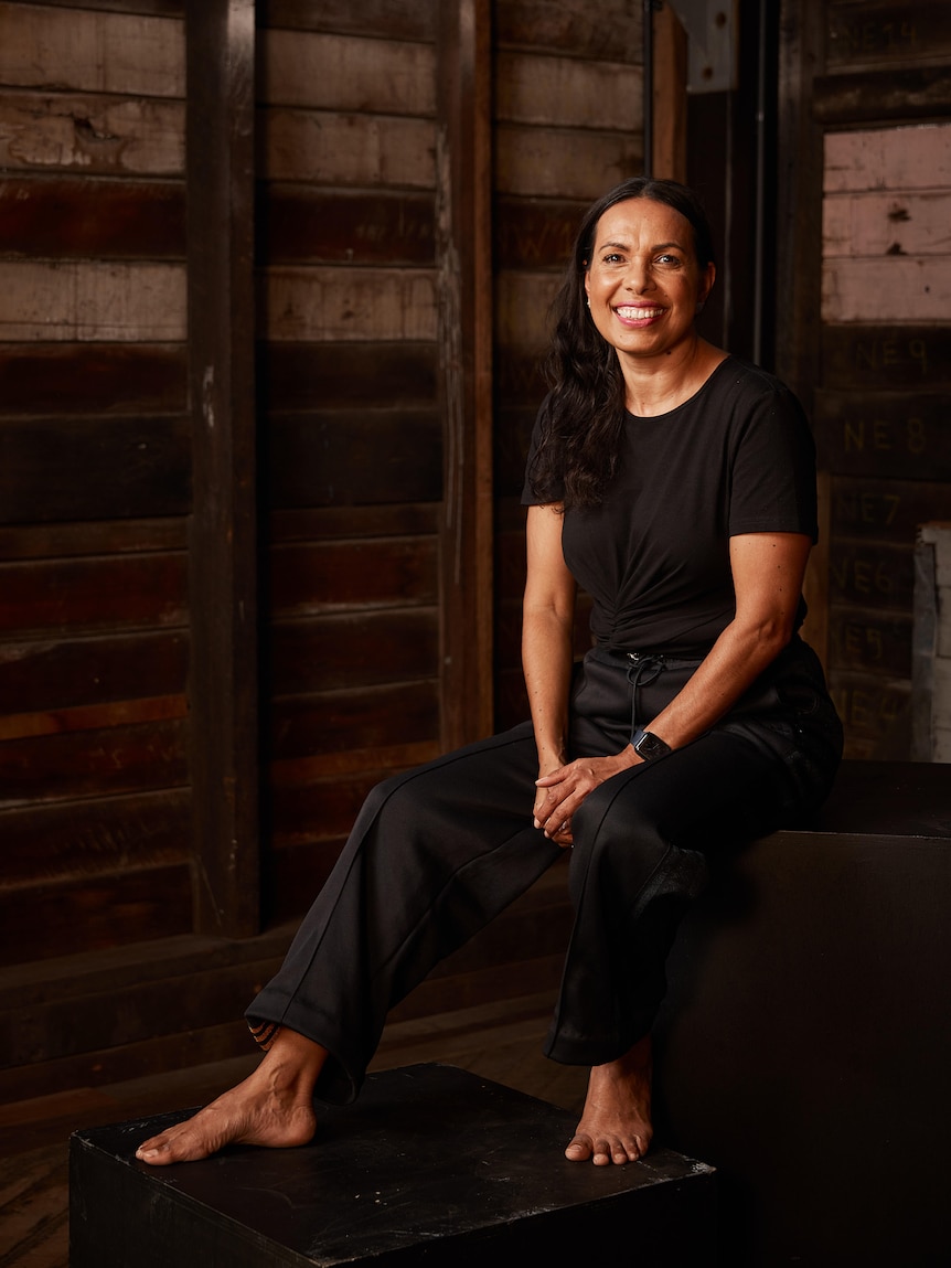 An Indigenous woman in her late 40s wearing black tshirt and pants and sitting in a wooden room, smiling