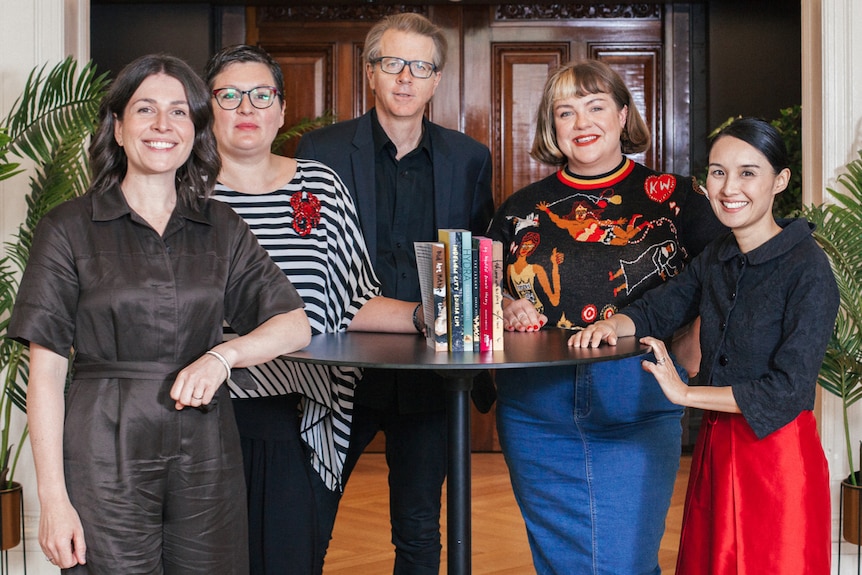 Five people - one man and four women - of varying ages and backgrounds stand around a table, on which six books are upright.