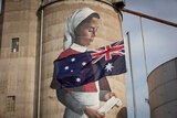 An Australian flag blows in the foreground of a large grain silo that has been painted with the image of a WWI nurse in uniform