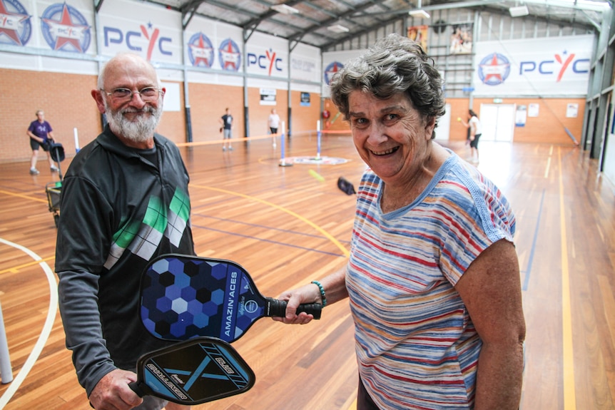 A smiling elderly  couple touch bats on an indoor sports court