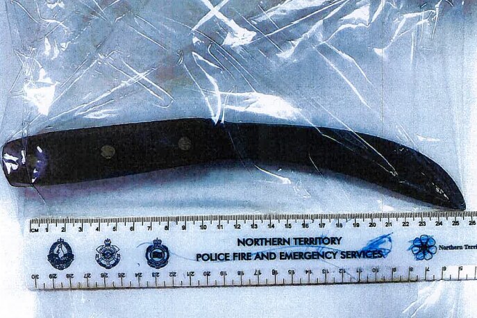 A large knife wrapped in a clear plastic bag, with a ruler running underneath it, lying on a flat surface. 