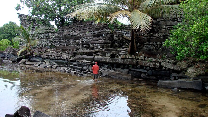 A man in a red jacket walks through a shallow pool towards a ruined wall.
