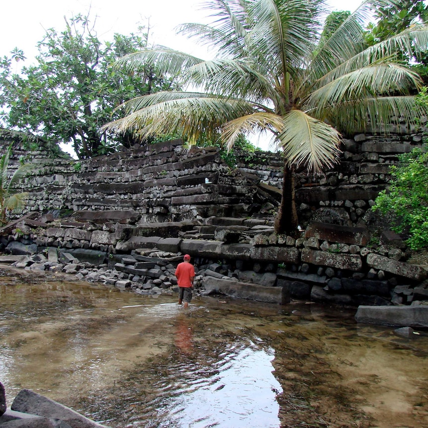 A man in a red jacket walks through a shallow pool towards a ruined wall.