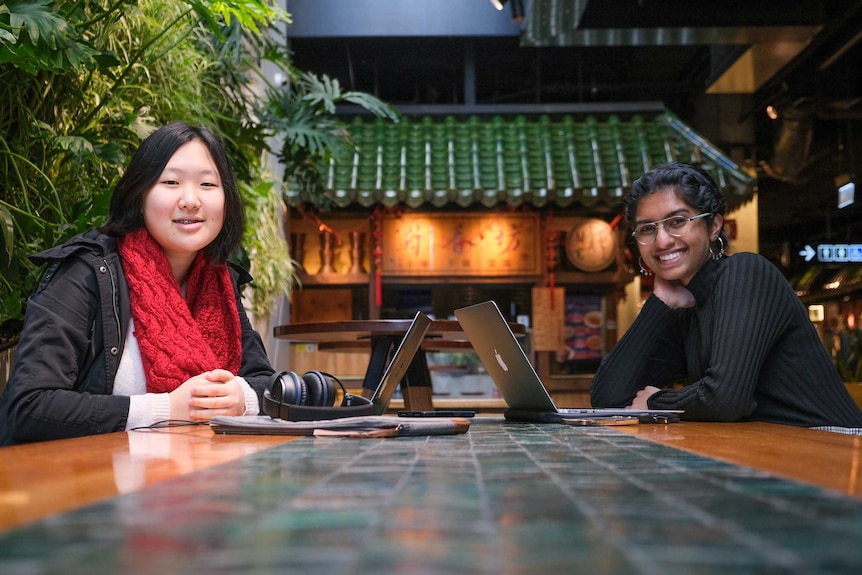 Hannah Ahn and Bagavathy staring into the camera while sitting across from each other in a food court.