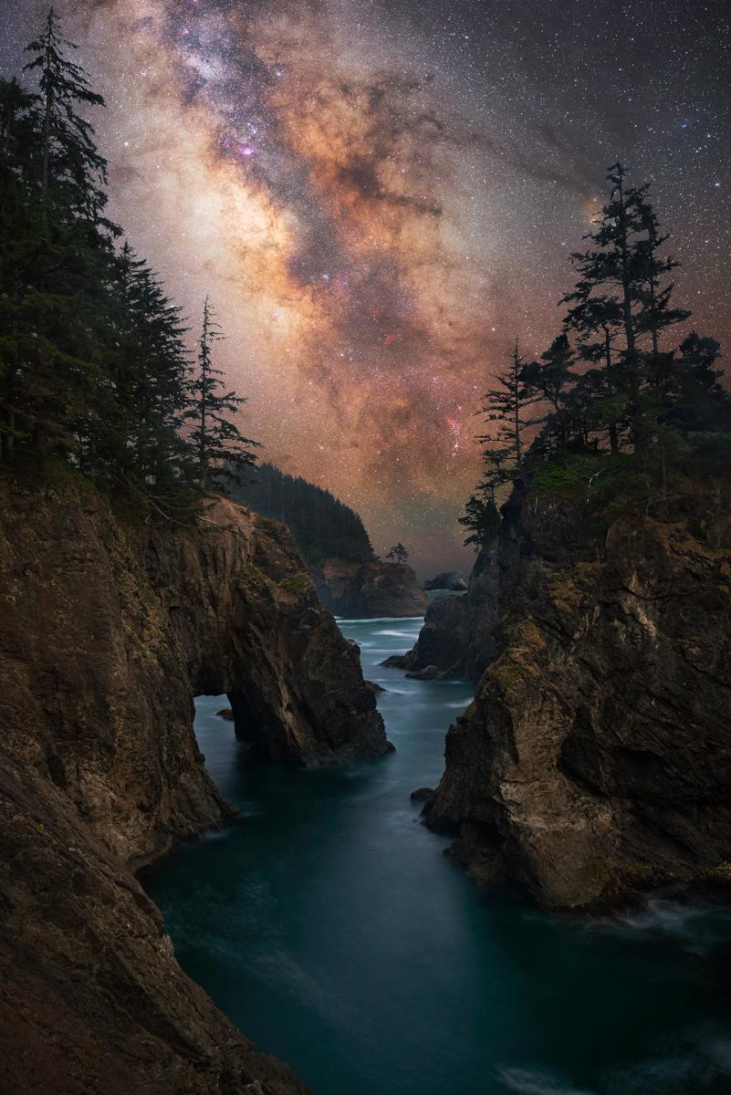 Milky Way seen between rows of trees and rocks over a river. 