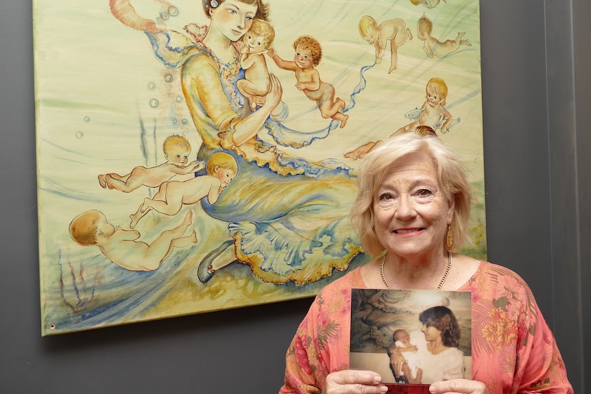 An older woman stands in front a a painting of a woman surrounded by babies, also holding a photo of her own baby.