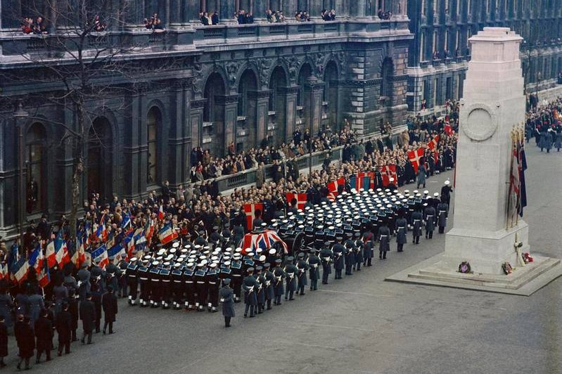 A coffin draped in the British flag is surrounded by naval officers in uniform.