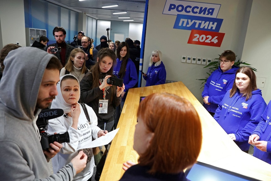 A large crowd of people stand at a desk, while several people wearing Putin campaign T-shirts look at them