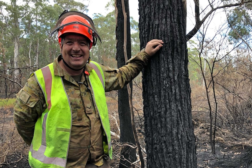 A smiling man in a high-visibility vest and red helmet standing next to a burnt tree.