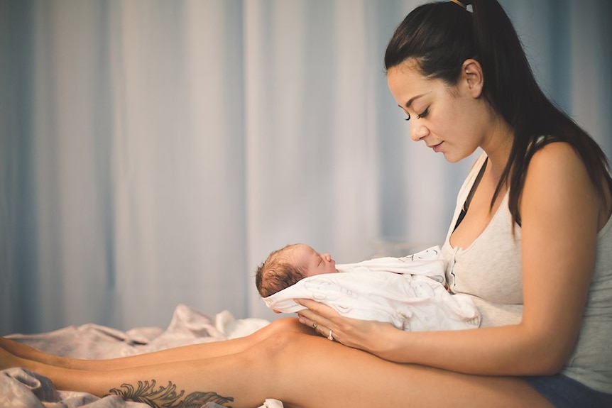 A mother looks down at her newborn who is lying on her legs wrapped in white cloth.