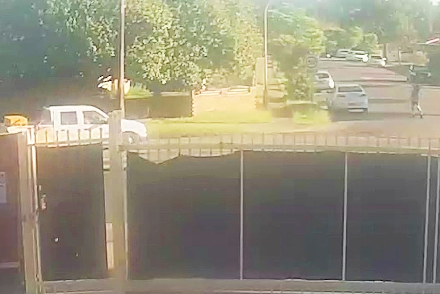 a cctv image of a white ute driving around a bend while a person crosses the street