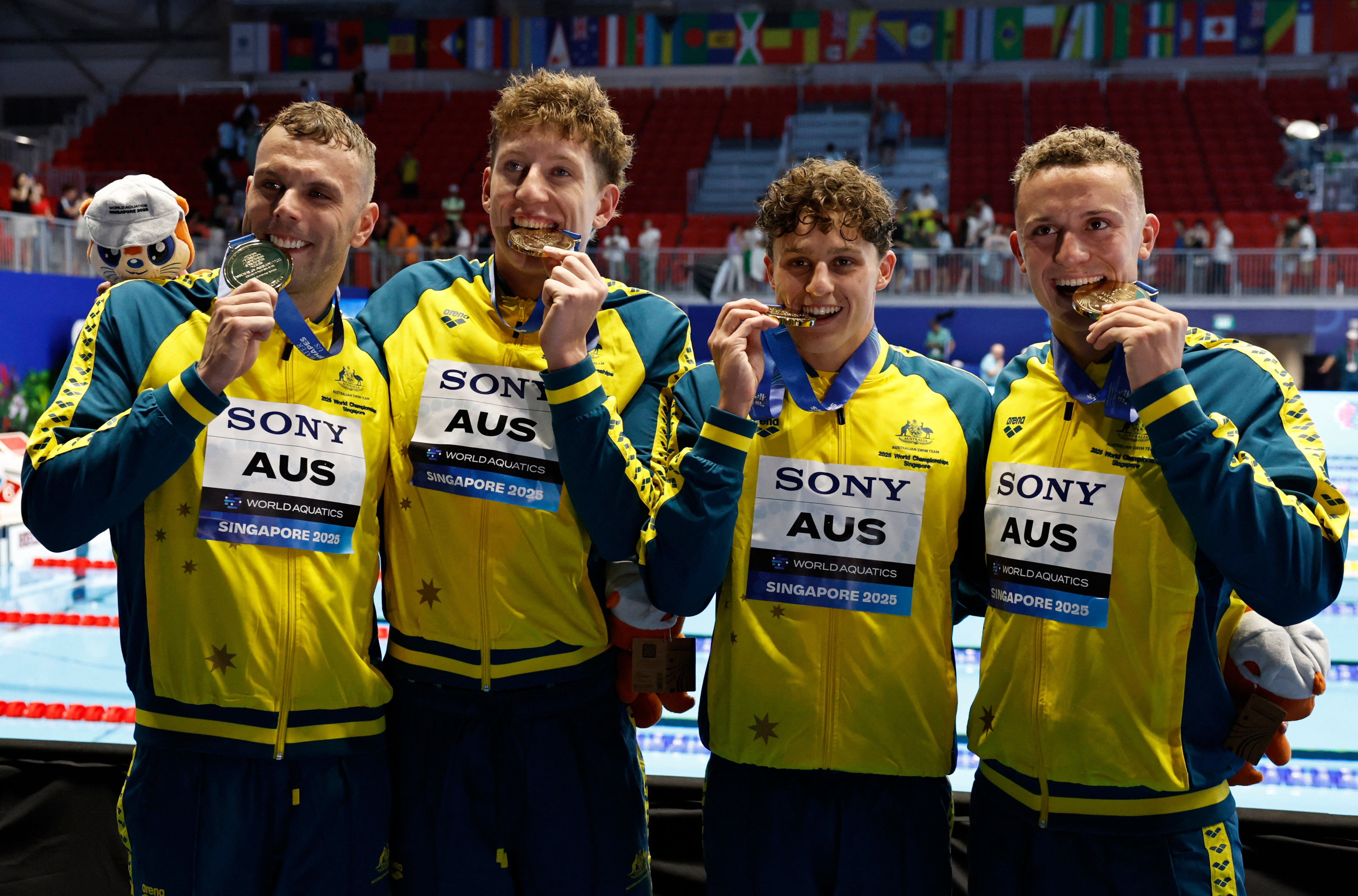 Four Australian swimmers holding gold medals by a large Olympic swimming pool.