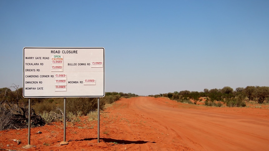 A road closure sign on red dirt and blue skies says the road to Warri Gate is open. 