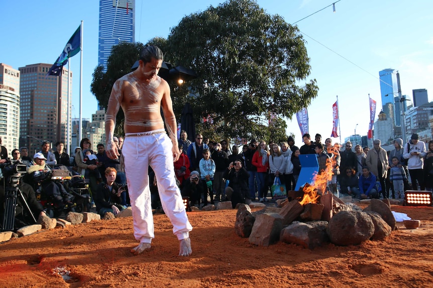 An Indigenous man stands by a fire at Federation Square, while people watch on.