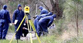 Police forensics officers in blue jumpsuits in bushland with a yellow tripod in the foreground.