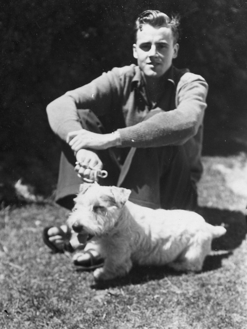 A black and white image of a young man sitting on the ground with a small white dog.