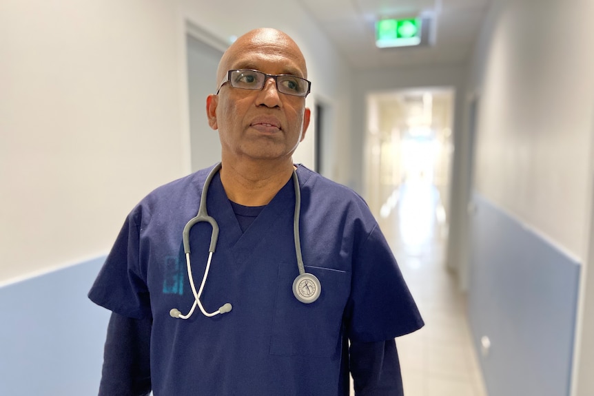 A man in scrubs with a stethoscope around his neck walks down a hallway 
