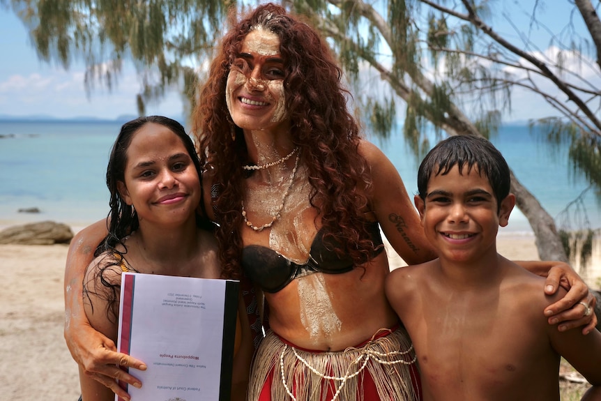 A painted Indigenous woman stands smiling between two Indigenous kids on an island holding a certificate