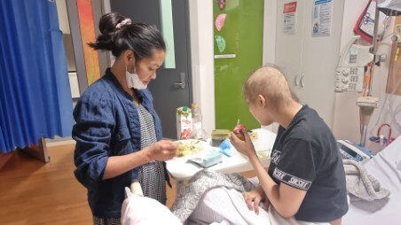 A woman stands next to a teenage girl in a hospital bed whose hair has fallen out from cancer treatment