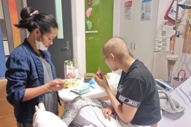 A woman stands next to a teenage girl in a hospital bed whose hair has fallen out from cancer treatment