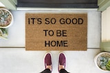 A person stands in front of a welcome mat that says "it's so good to be home".