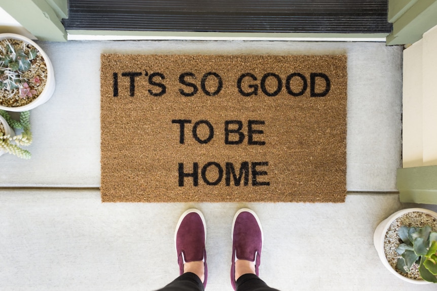 A person stands in front of a welcome mat that says "it's so good to be home".