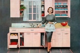 A grainy, old-looking coloured image of a woman in 1950s-style dress and apron, standing smiling near a sink in a tidy kitchen.