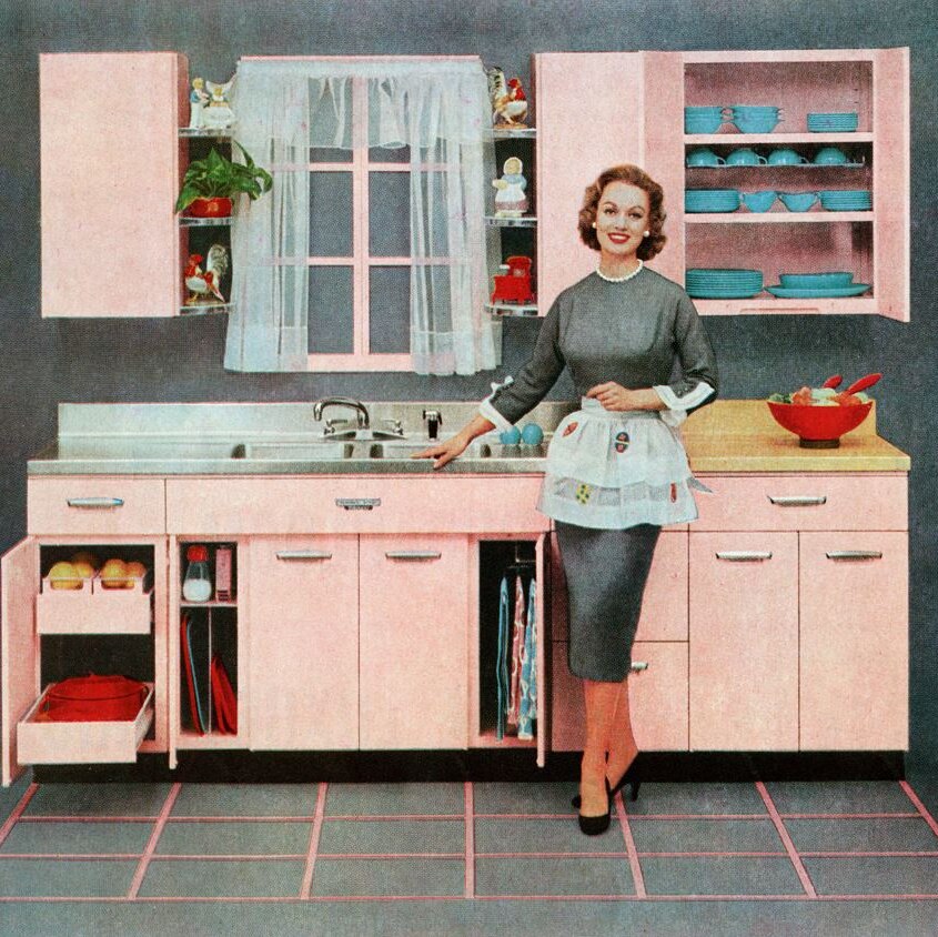 A grainy, old-looking coloured image of a woman in 1950s-style dress and apron, standing smiling near a sink in a tidy kitchen.