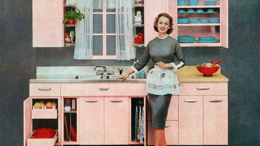 A grainy, old-looking coloured image of a woman in 1950s-style dress and apron, standing smiling near a sink in a tidy kitchen.