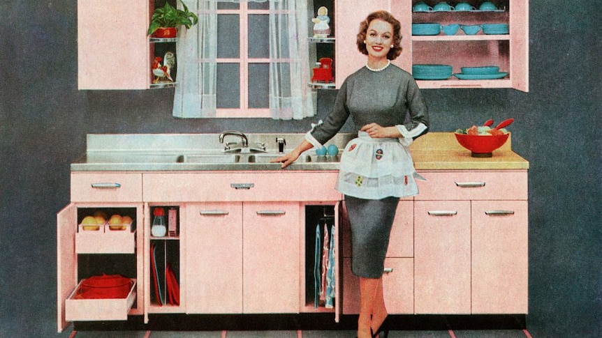 A grainy, old-looking coloured image of a woman in 1950s-style dress and apron, standing smiling near a sink in a tidy kitchen.