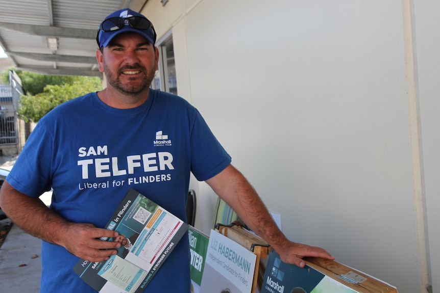 Man with blue hat and shirt on smiling holding ballot papers.