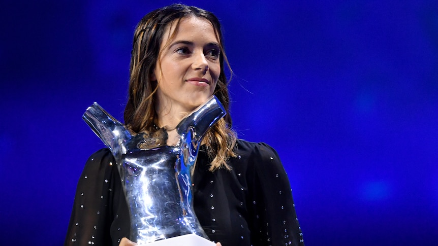 A star Spanish women's footballer looks off to the side of stage as she stands holding a trophy.