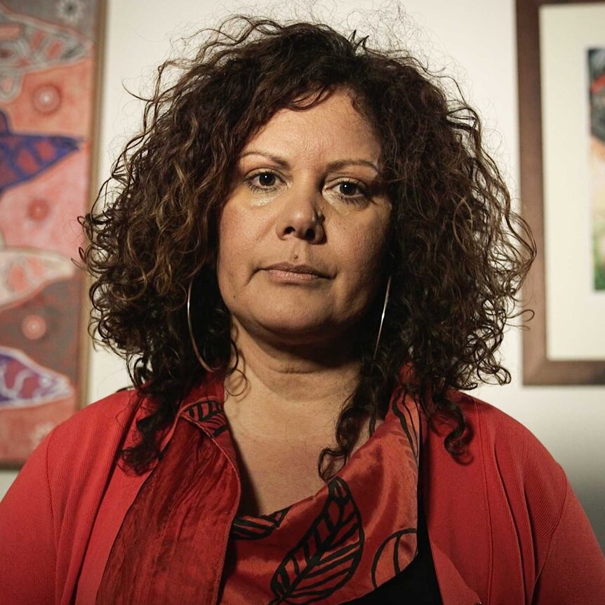 Malarndirri McCarthy, wearing red, looks straight down the barrel. Aboriginal art is visible on the walls behind her.