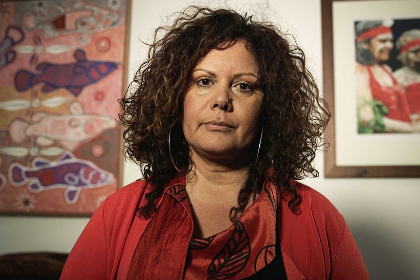 Malarndirri McCarthy, wearing red, looks straight down the barrel. Aboriginal art is visible on the walls behind her.