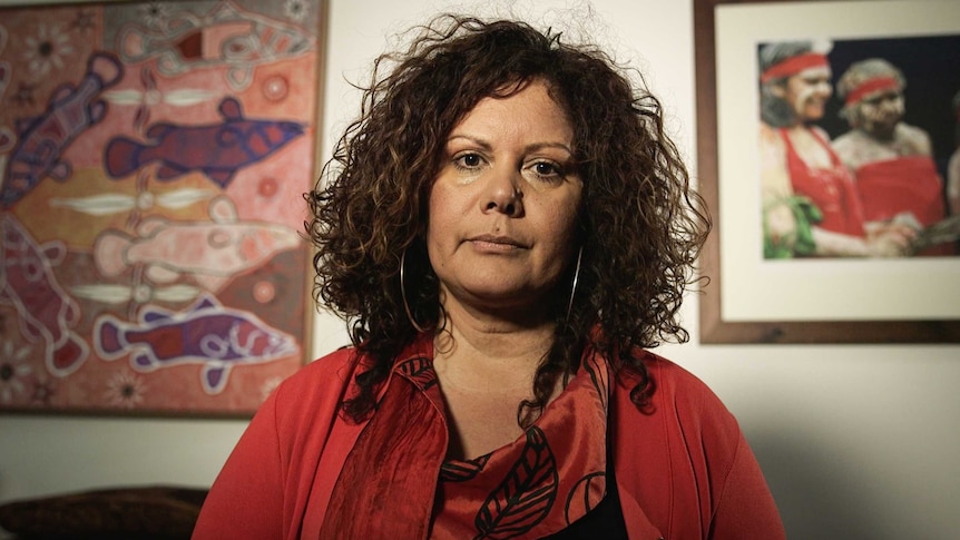 Malarndirri McCarthy, wearing red, looks straight down the barrel. Aboriginal art is visible on the walls behind her.