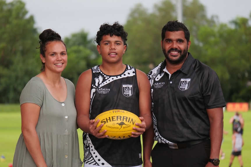 A boy smiles while holding a football, flanked on either side by his parents.