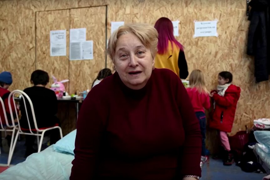 An older woman with short blonde hair wearing a mauve jumper sits on a makeshift bed, looking at the camera