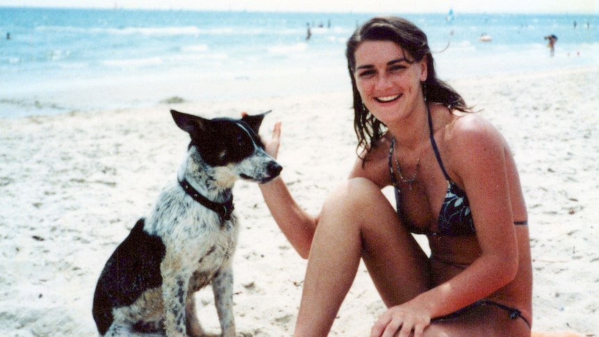 A young woman and her dog sit next to each other on a beach.