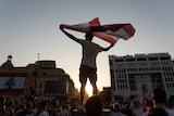 A silhouette against the sky of a man holding a flag above a crowd of people.   
