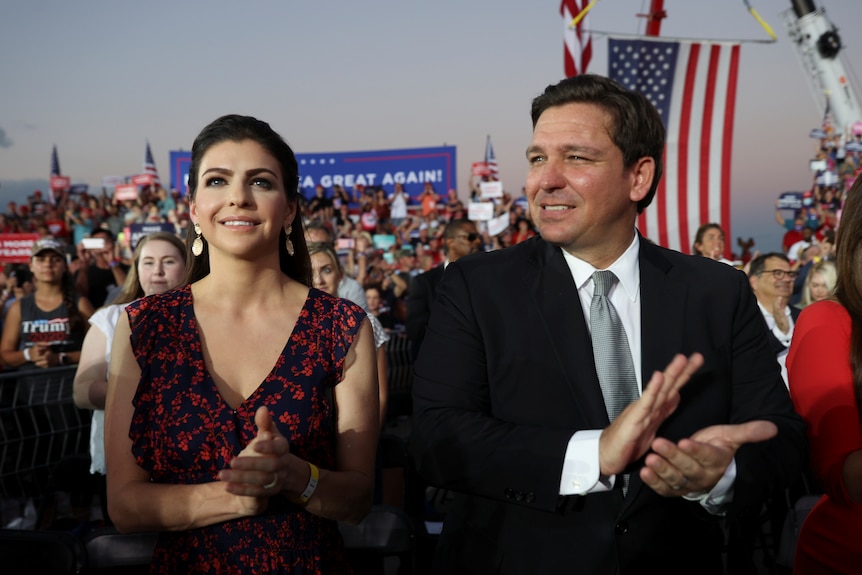 Ron Desantis, in a black suit, stands next to his wife who is brunette and wearing a black-and-red dress