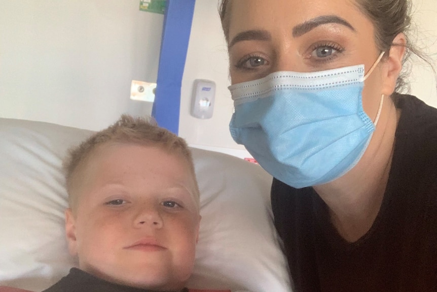 A woman wearing a face mask takes a selfie with her eight-year-old son who is lying on a hospital bed