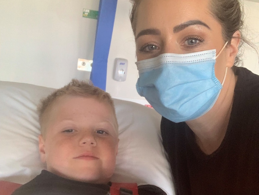 A woman wearing a face mask takes a selfie with her eight-year-old son who is lying on a hospital bed