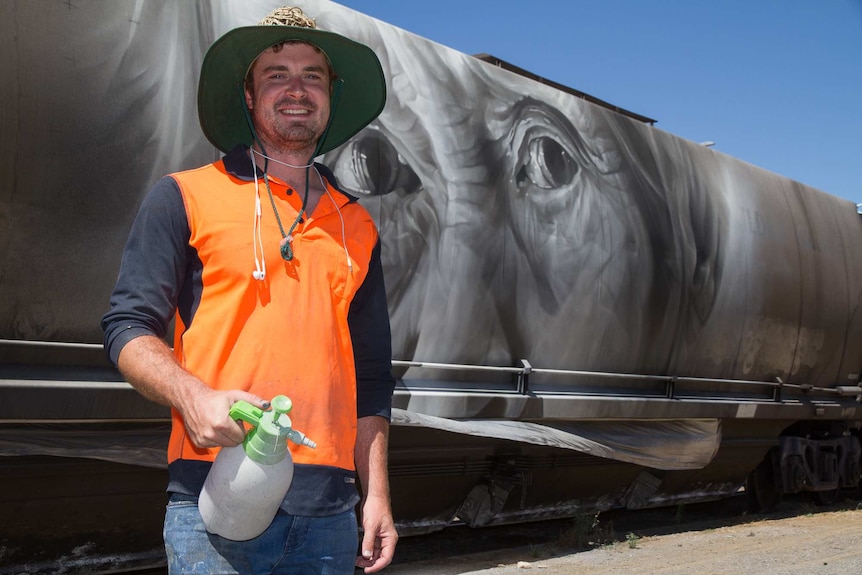 A man in high visibility gear holding a spray bottle standing in front of a rail carriage with a face painted on it