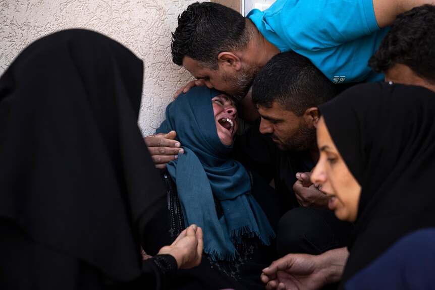 A woman in blue headscarf cries as man kisses her forehead and other people surround her. 