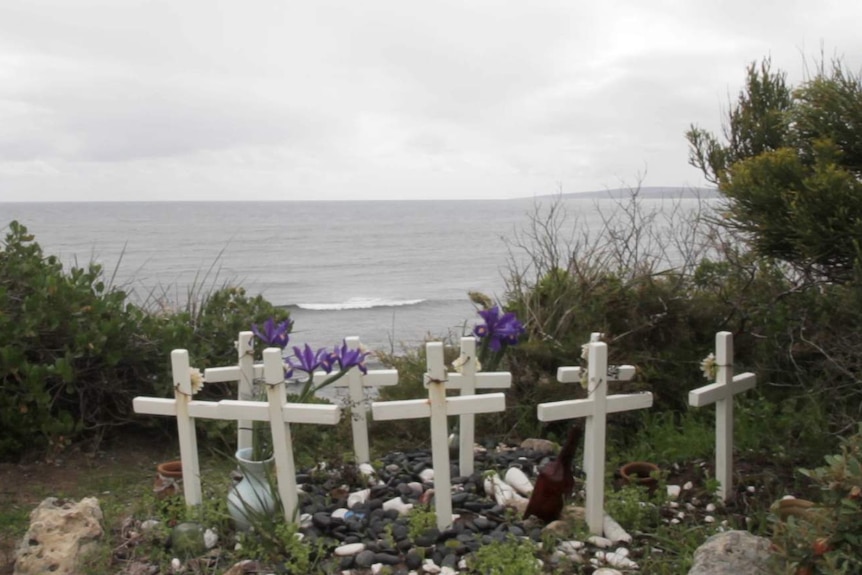 Nine white crosses adorned with flowers are erected in a circle on the beach above the cliff.