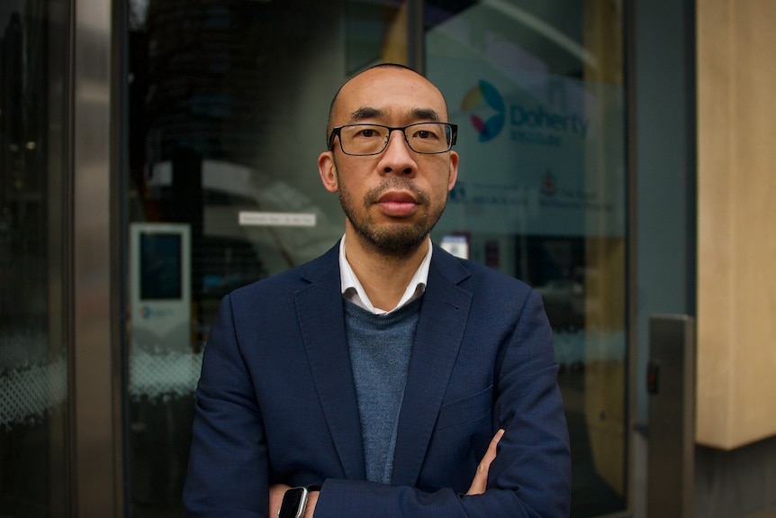 A balding, Asian man in glasses wearing a suit jacket over a woollen jumper and white shirt stands in front of a building.
