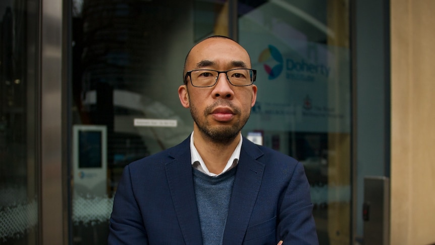 A man in glasses wearing a suit jacket over a woollen jumper stands in front of a building.