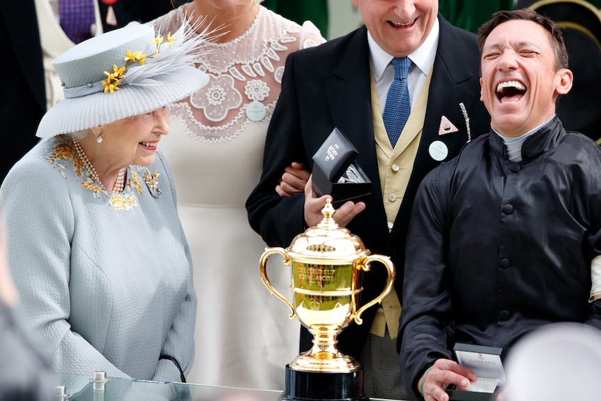 A jockey laughs as he stands opposite Queen Elizabeth II after a big race, with a gold trophy between them. 