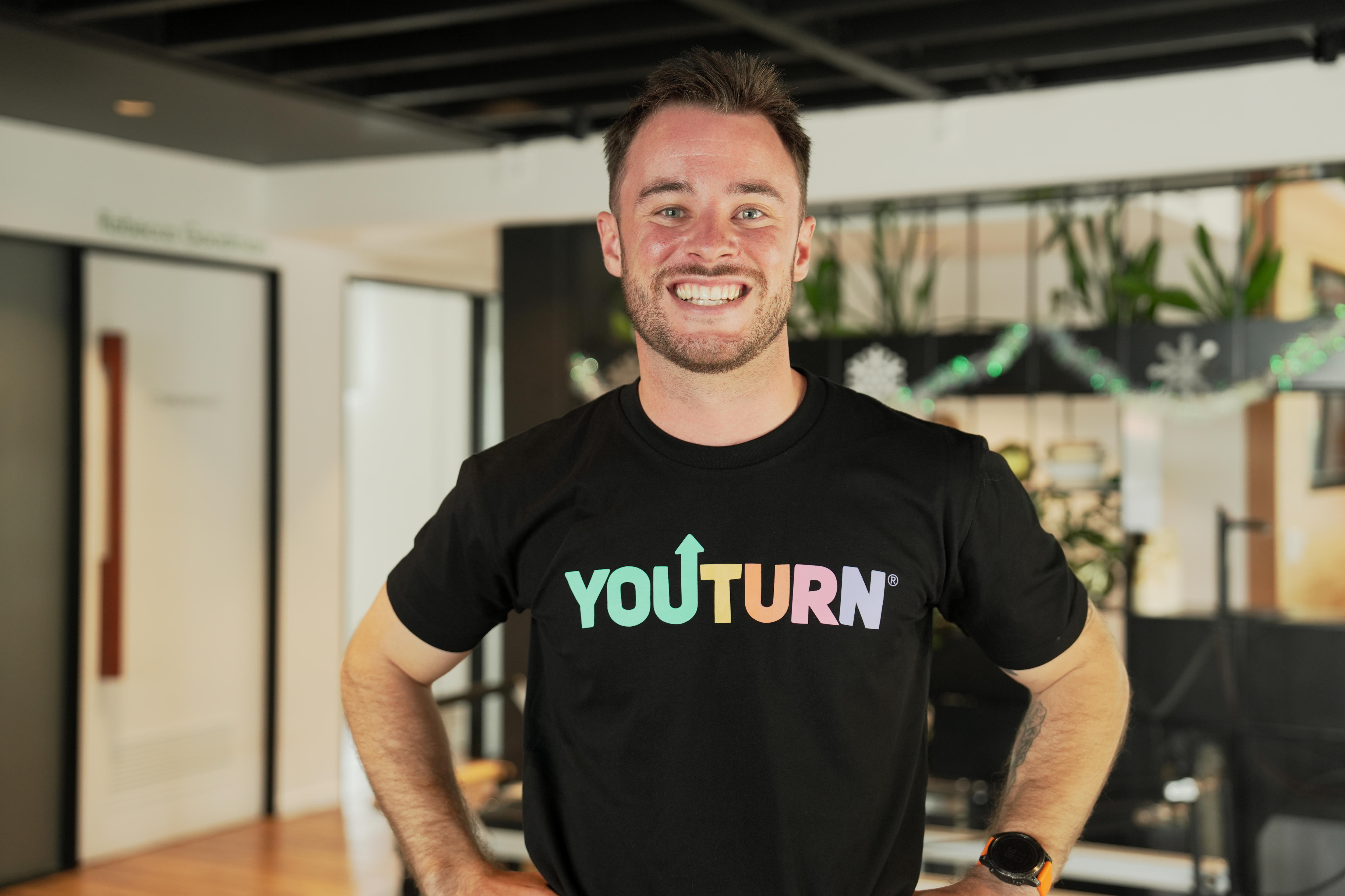     Un hombre con una camiseta negra lee "tu giras" sonriendo a la cámara en un gimnasio