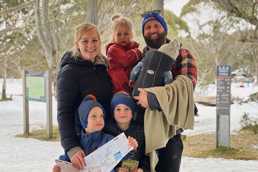A man in a beanie stands smiling with his wife and four children, surrounded by snow.